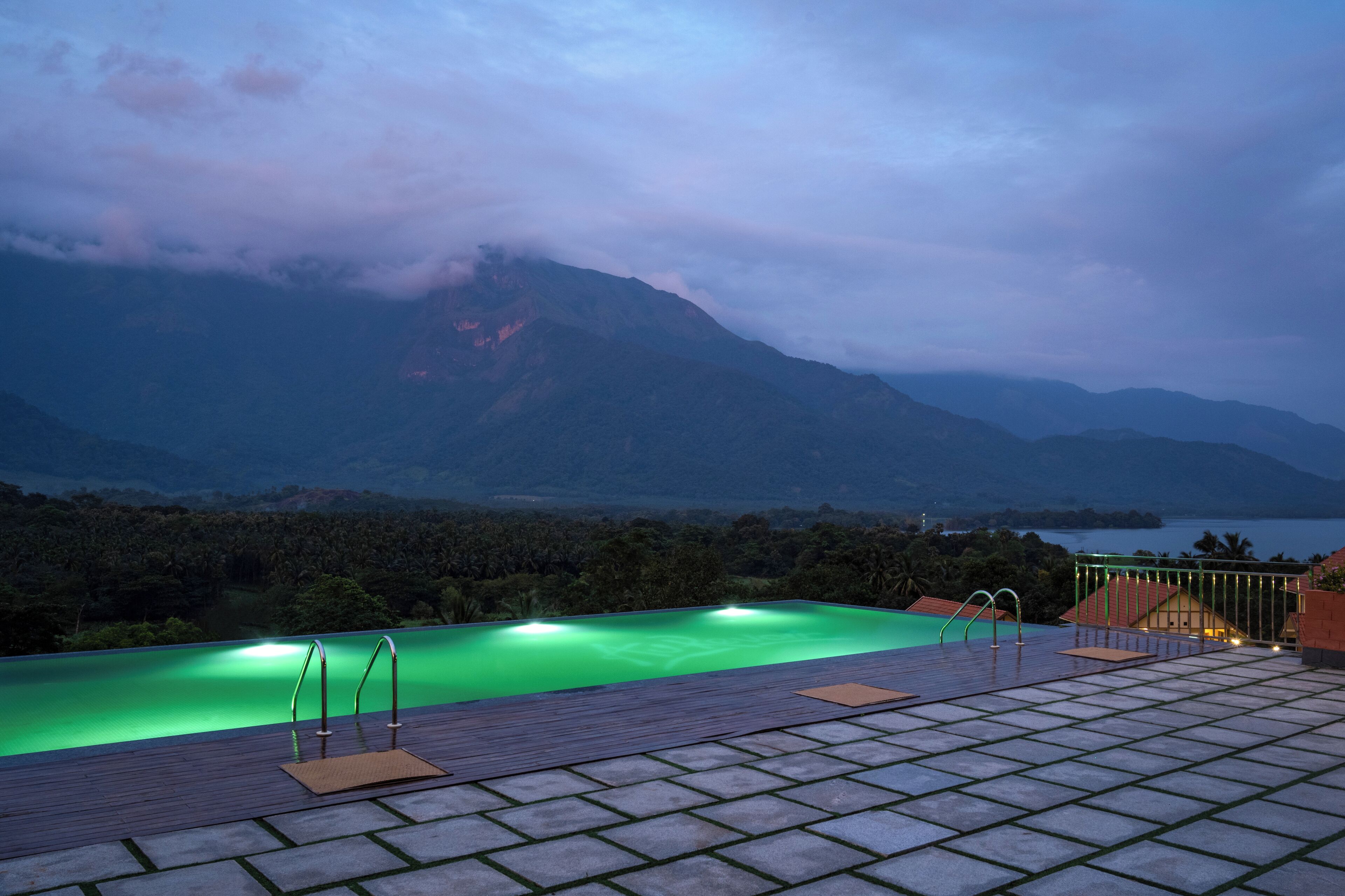 A rooftop infinity pool with a view of mountains and a lake at dusk.