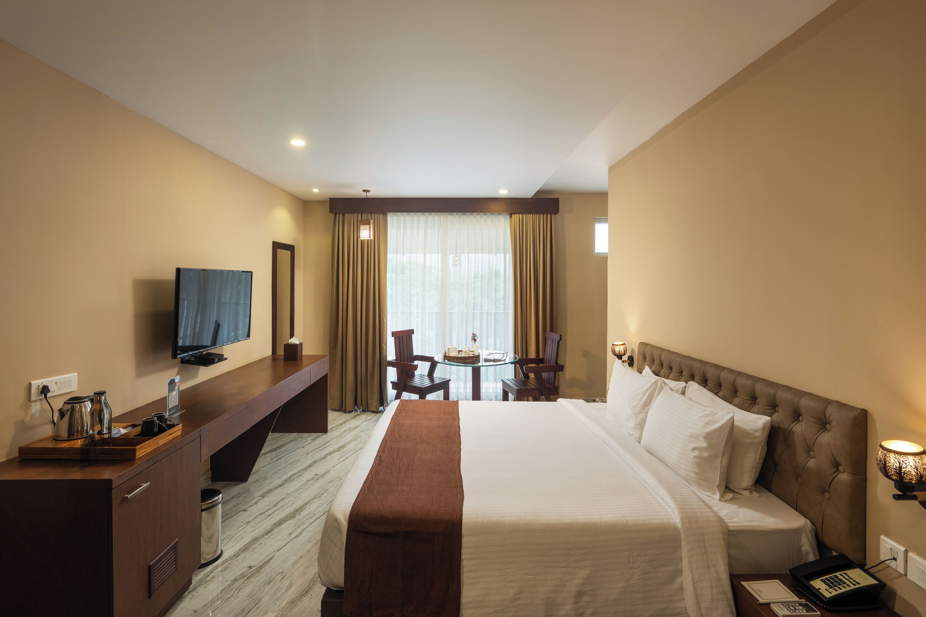 A hotel room with a bed, a TV, a desk with a lamp, a chair, and a small table with a cake.