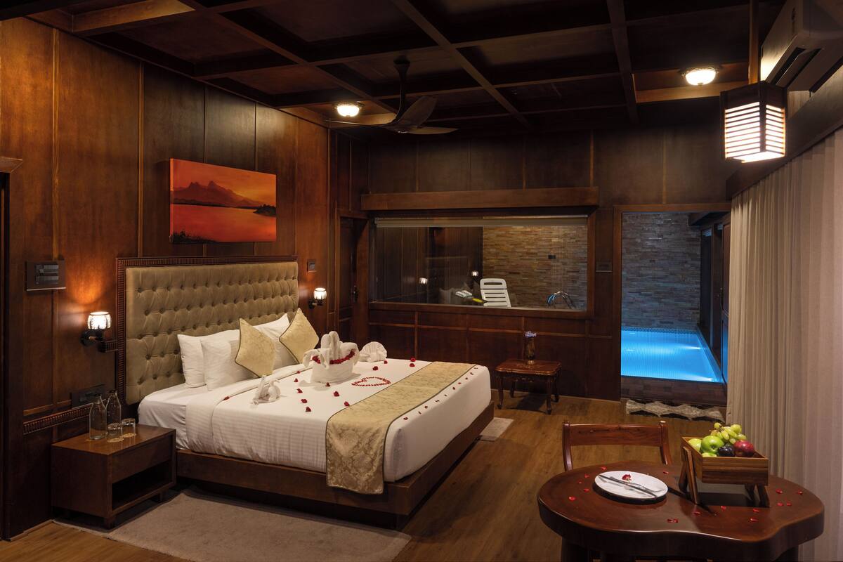 Une chambre spacieuse avec un grand lit, une tête de lit en bois, une table de chevet, une petite table avec un panier de fruits et une vue sur la piscine par une grande fenêtre.