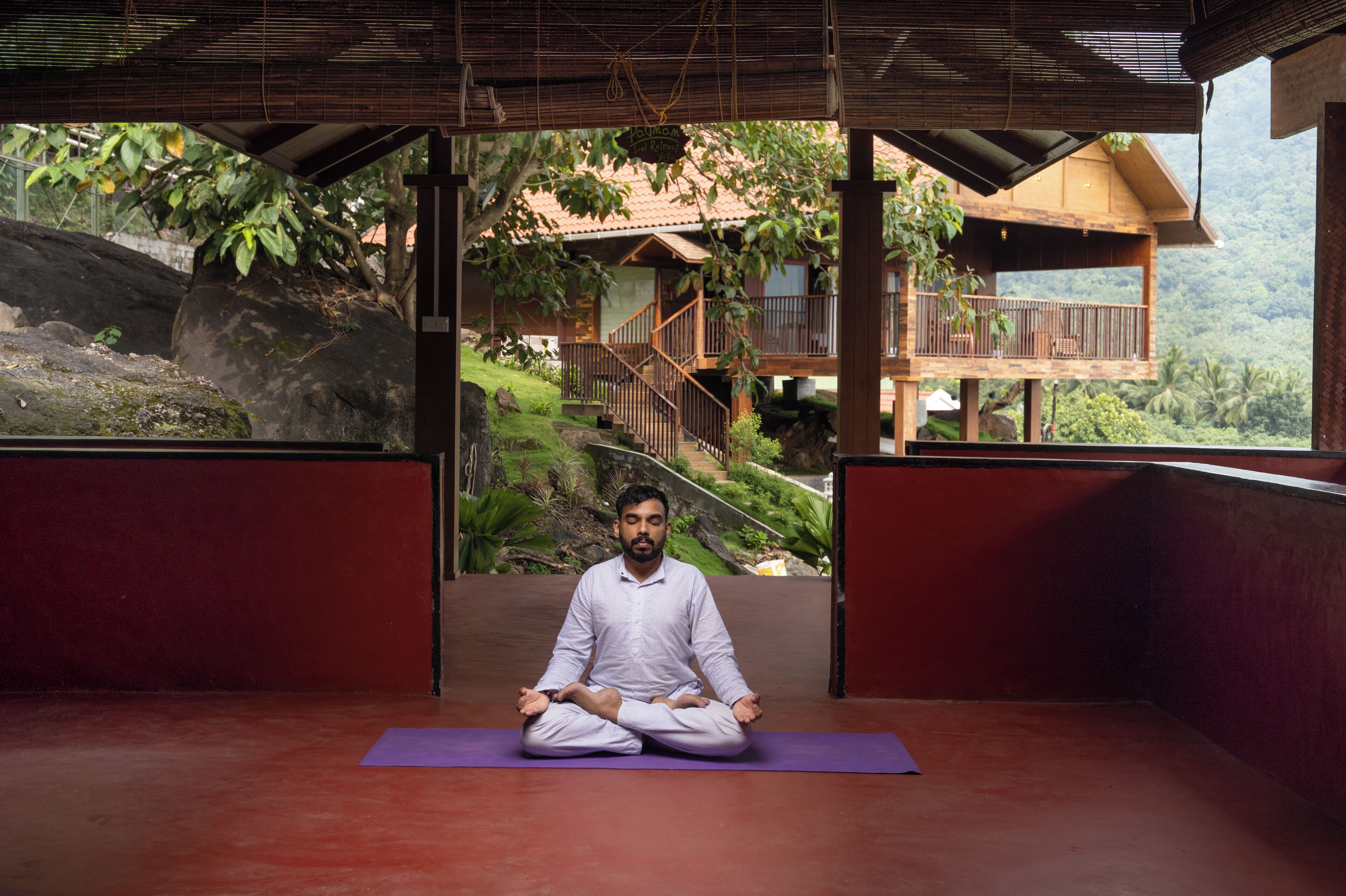 A person meditating on a purple mat in a room with a view of a wooden structure and greenery.