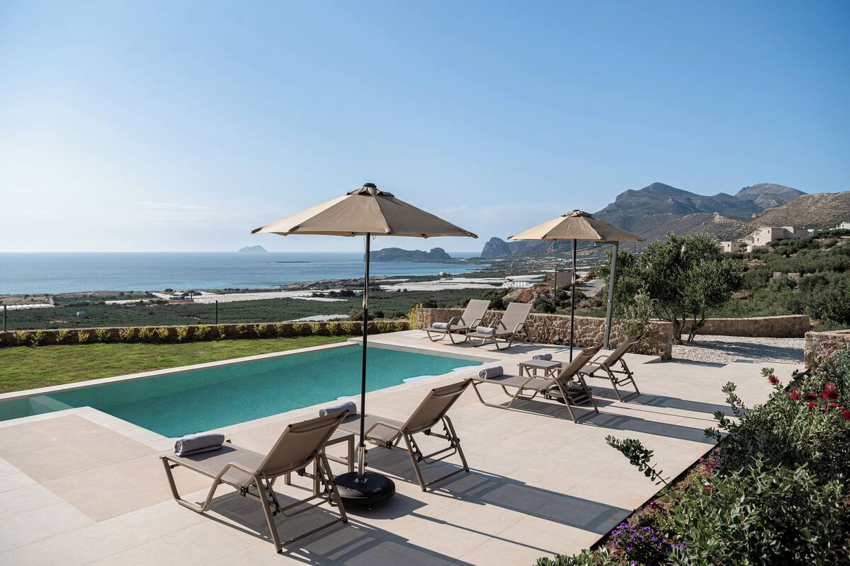 A poolside area with lounge chairs and umbrellas overlooking a clear blue sea and distant mountains.