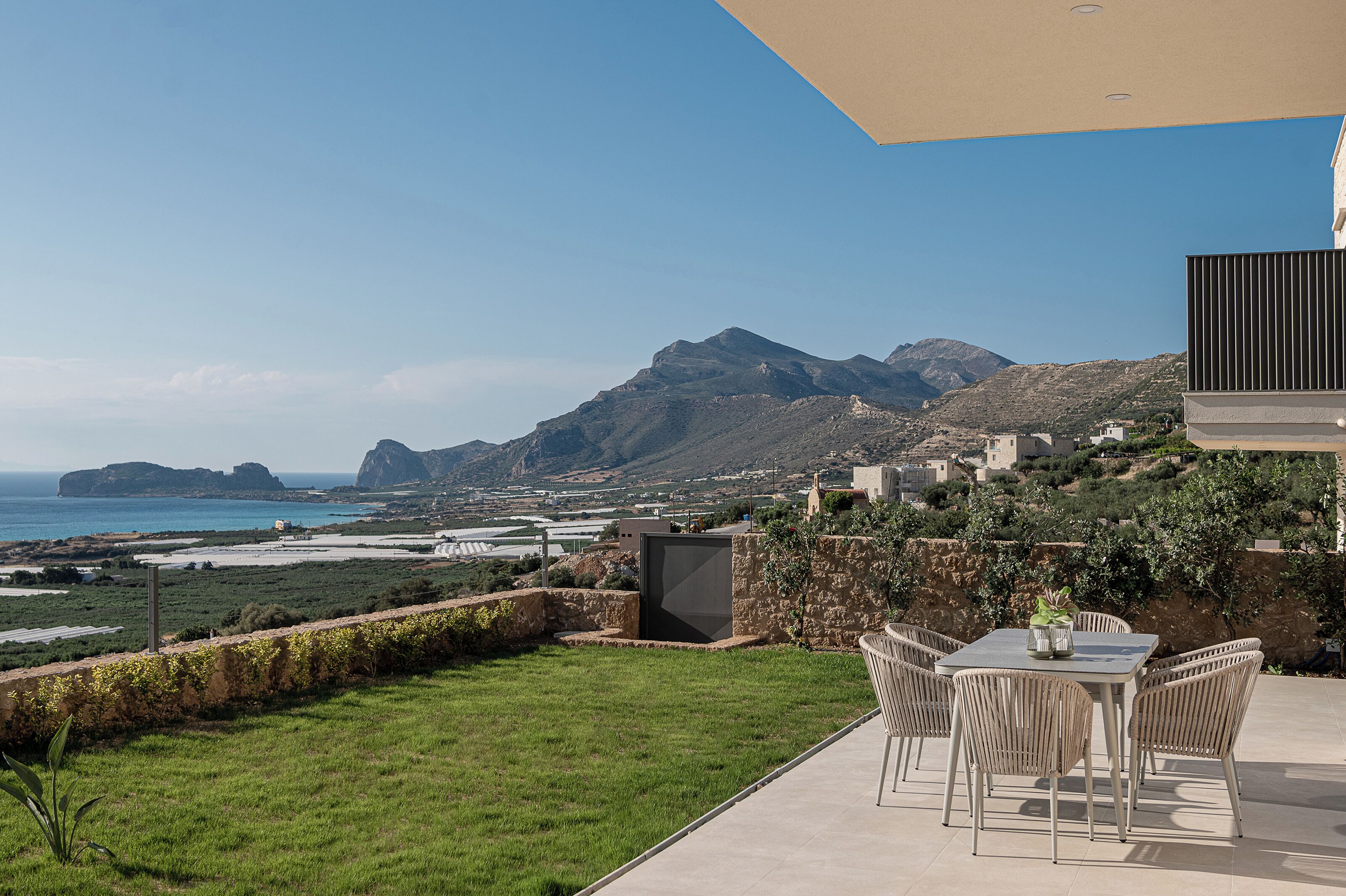 Eine Terrasse mit Tisch und Stühlen mit Blick auf eine Küstenlandschaft, im Hintergrund Berge.