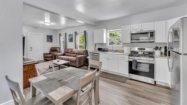 A modern kitchen with white cabinets, stainless steel appliances, and a dining area with a wooden table and chairs. - Pitcher's Paradise Apt 1 - All Star Baseball Rentals (Oneonta)