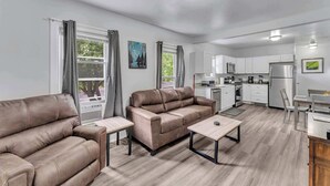 A modern living room with a sofa, coffee table, and dining area. It includes a kitchen with white cabinets and appliances. - Pitcher's Paradise Apt 1 - All Star Baseball Rentals (Oneonta)