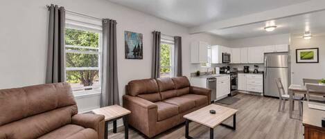A modern living room with two sofas, a coffee table, and a kitchen area with white cabinets and appliances.