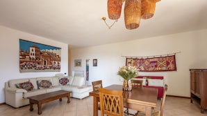A living room with a sofa, a wooden dining table, and chairs. There is a painting on the wall and a tapestry.