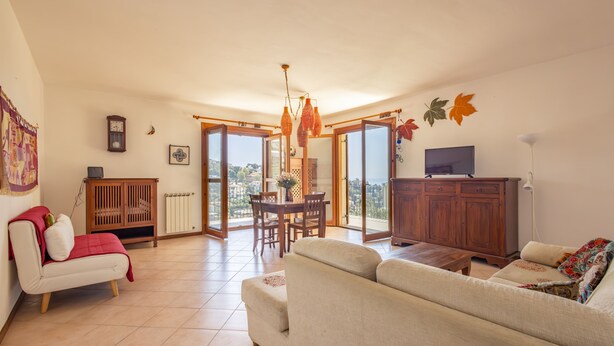 A spacious living room with a sofa, a dining table, and a TV. There is a view through the glass doors to the outside.