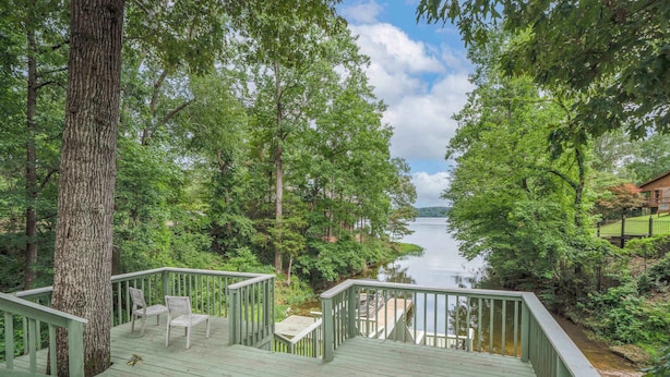 A wooden deck with chairs and a table, overlooking a lake surrounded by trees.