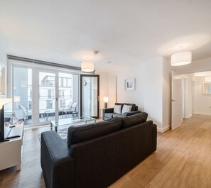 Una sala moderna con un sofá gris oscuro, una mesa de centro de cristal y vistas a un balcón con mobiliario exterior.