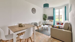 A modern living room with a wooden dining table, white chairs, a beige sofa, and a TV.
