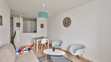 Ein modernes Wohnzimmer mit einer Couch, einem Couchtisch, zwei Sesseln, einem Esstisch mit Stühlen, einer Pendelleuchte und einer Wanduhr.