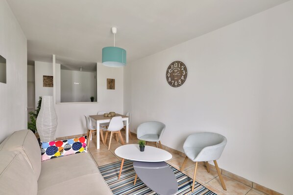 A modern living room with a sofa, a coffee table, two chairs, a dining table with chairs, a hanging light fixture, and a wall clock. - 123HOME - PARK SIDE (Chessy)