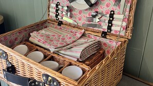 A wicker picnic basket with a floral pattern, containing neatly folded napkins, a plate, cutlery, and candles.