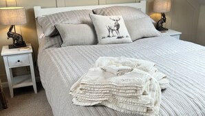 A neatly made bed with a quilted grey duvet, two pillows with a deer print, and a stack of folded towels. There are two bedside tables with lamps and a small statue of a rabbit on one.