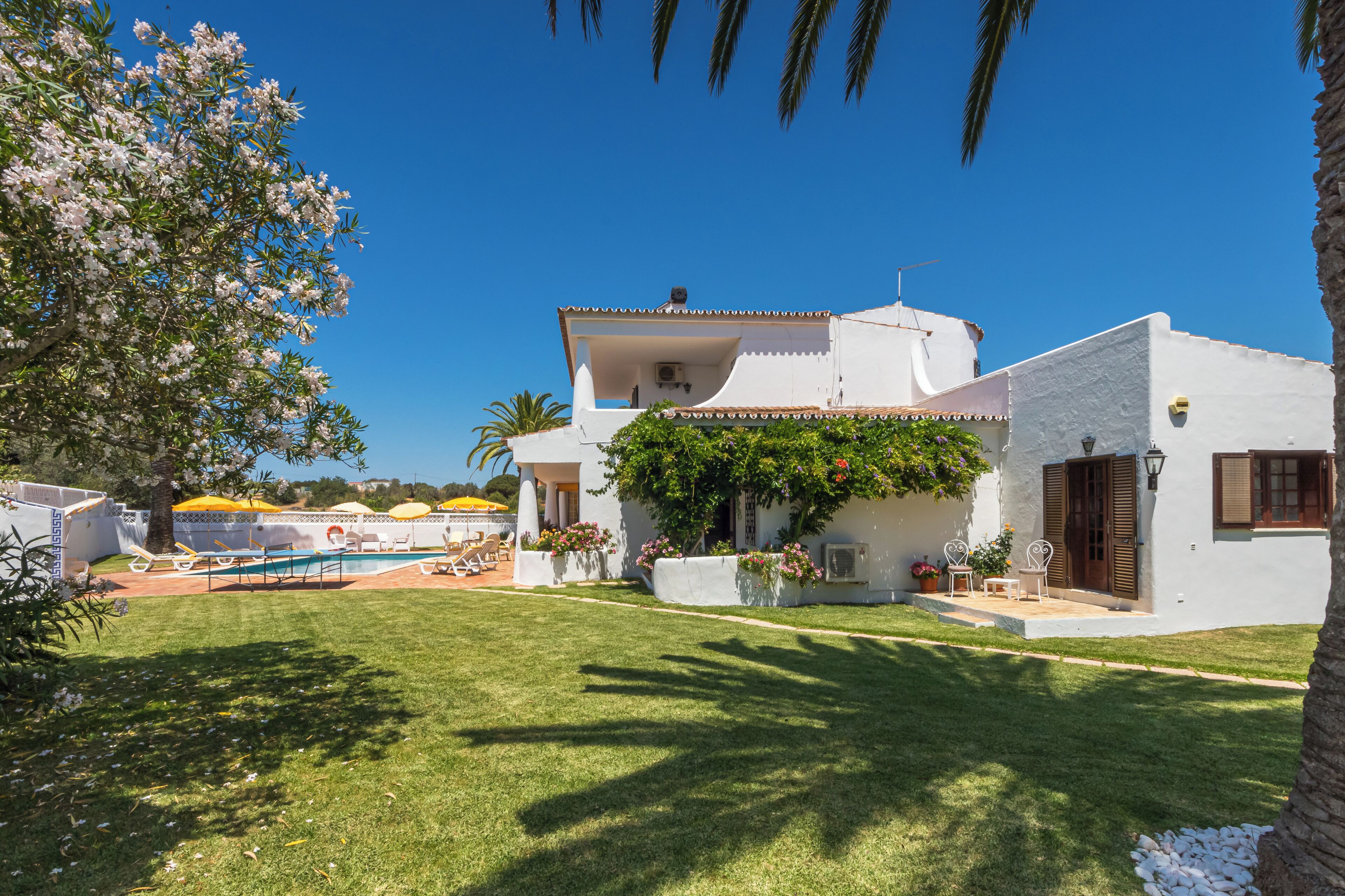 Una villa blanca con piscina, rodeada de vegetación y un cielo azul claro.
