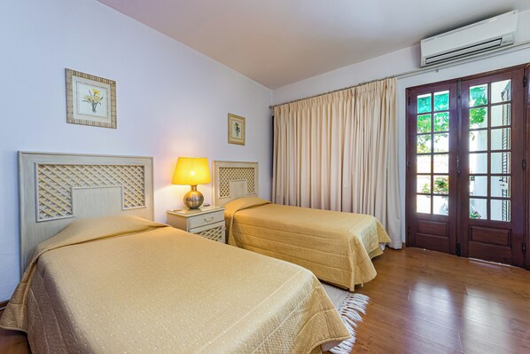 A hotel room with two beds, a nightstand with a lamp, a window with curtains, and an air conditioning unit.