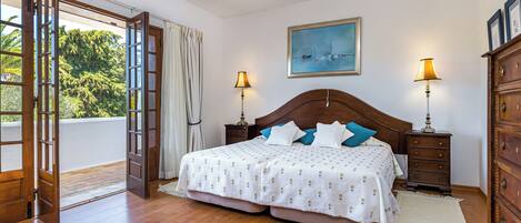 A bedroom with a large bed, wooden headboard, two bedside tables with lamps, a nightstand, a dresser, a painting on the wall, and a view of greenery through an open door.