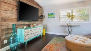 A living room with a wooden wall, a flat-screen TV, a teal dresser, a yellow trash bin, and a beige ottoman. - Spacious 3-BR Home with Patio, Firepit & BBQ! (Largo)