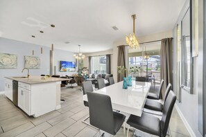A modern kitchen with a central island, dining area with a long table and chairs, and large windows with curtains.