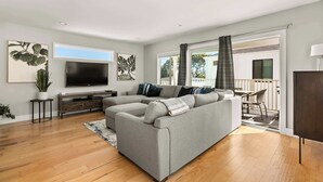 A modern living room with a grey sectional sofa, wooden flooring, a flat-screen TV, and a balcony with a white fence. - Modern Pacific Beach Paradise (San Diego)