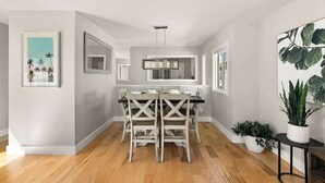A modern kitchen with a central island, white cabinetry, and a dining area with a white table and chairs. There are potted plants and a framed picture on the walls. - Modern Pacific Beach Paradise (San Diego)