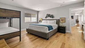 A modern bedroom with a bunk bed, a large bed with a grey frame, wooden flooring, a window with blinds, and a hallway leading to another room. - Modern Pacific Beach Paradise (San Diego)