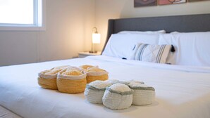 Cama bem arrumada com edredom branco e travesseiros, luminária de cabeceira e janela com luz natural.