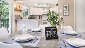 A modern dining area with a table set for two, a sign that reads "EAT DRINK ENJOY", and a view of the kitchen in the background. - Modern 2BR/2BA Kirkland Home, Walk to Beach Park (Kirkland)