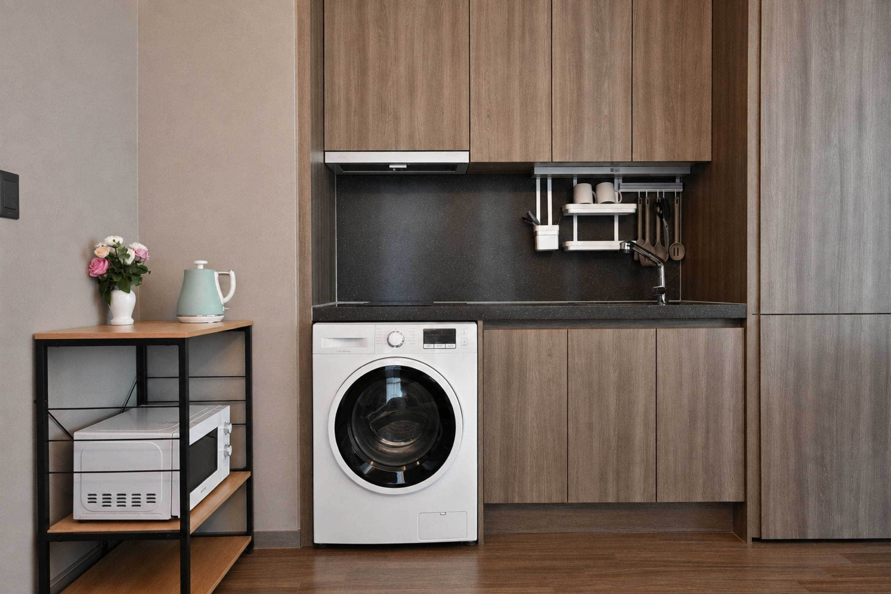 Cocina compacta con lavadora integrada, armarios de madera y un pequeño estante con un jarrón y una tetera.