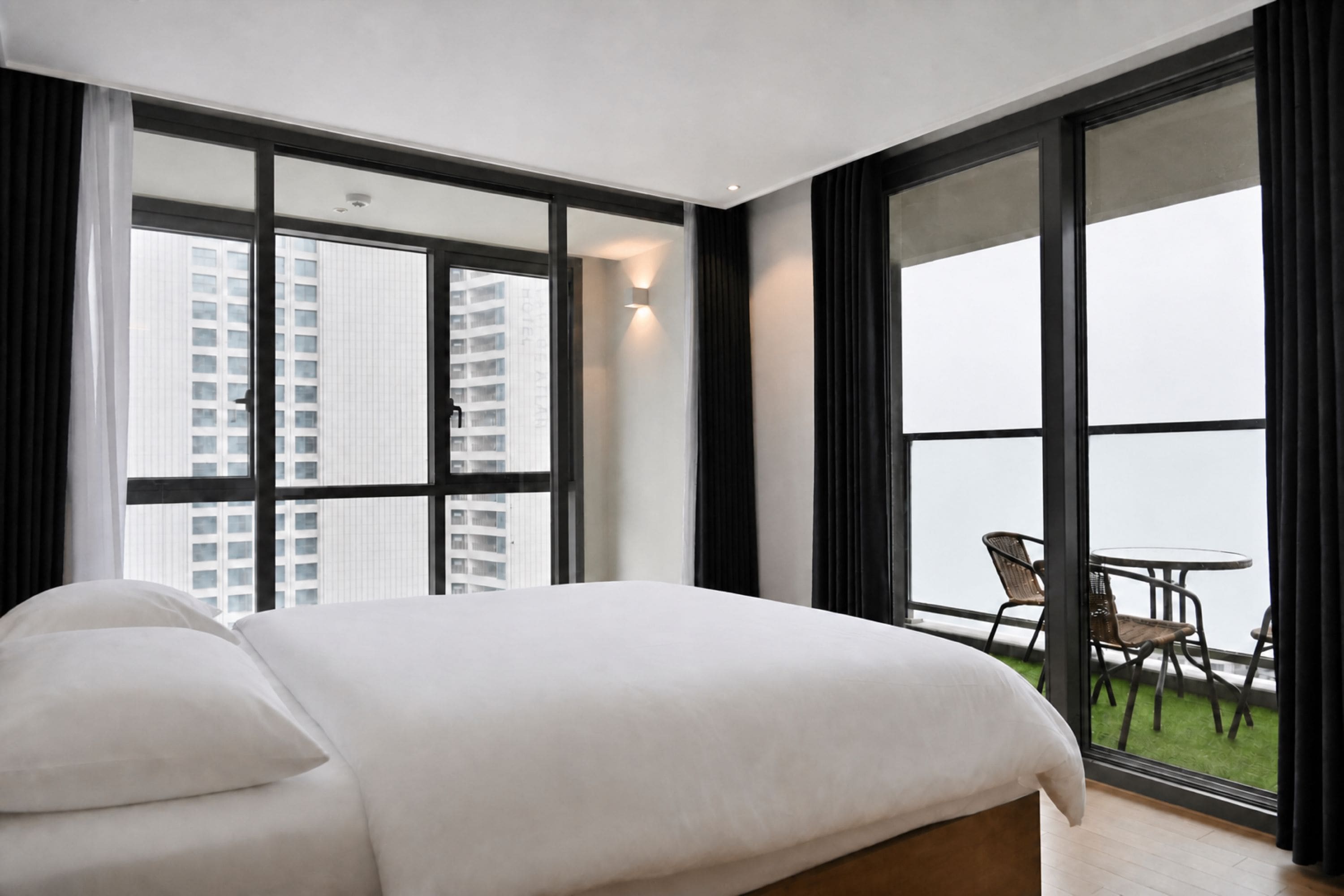 Una habitación de hotel moderna con una cama grande, un balcón con mesa y sillas, y vistas a edificios altos.