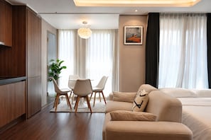 Una habitación de hotel moderna con un armario de madera, una mesa de comedor con sillas, un sofá, un ventanal con cortinas y un cuadro en la pared.