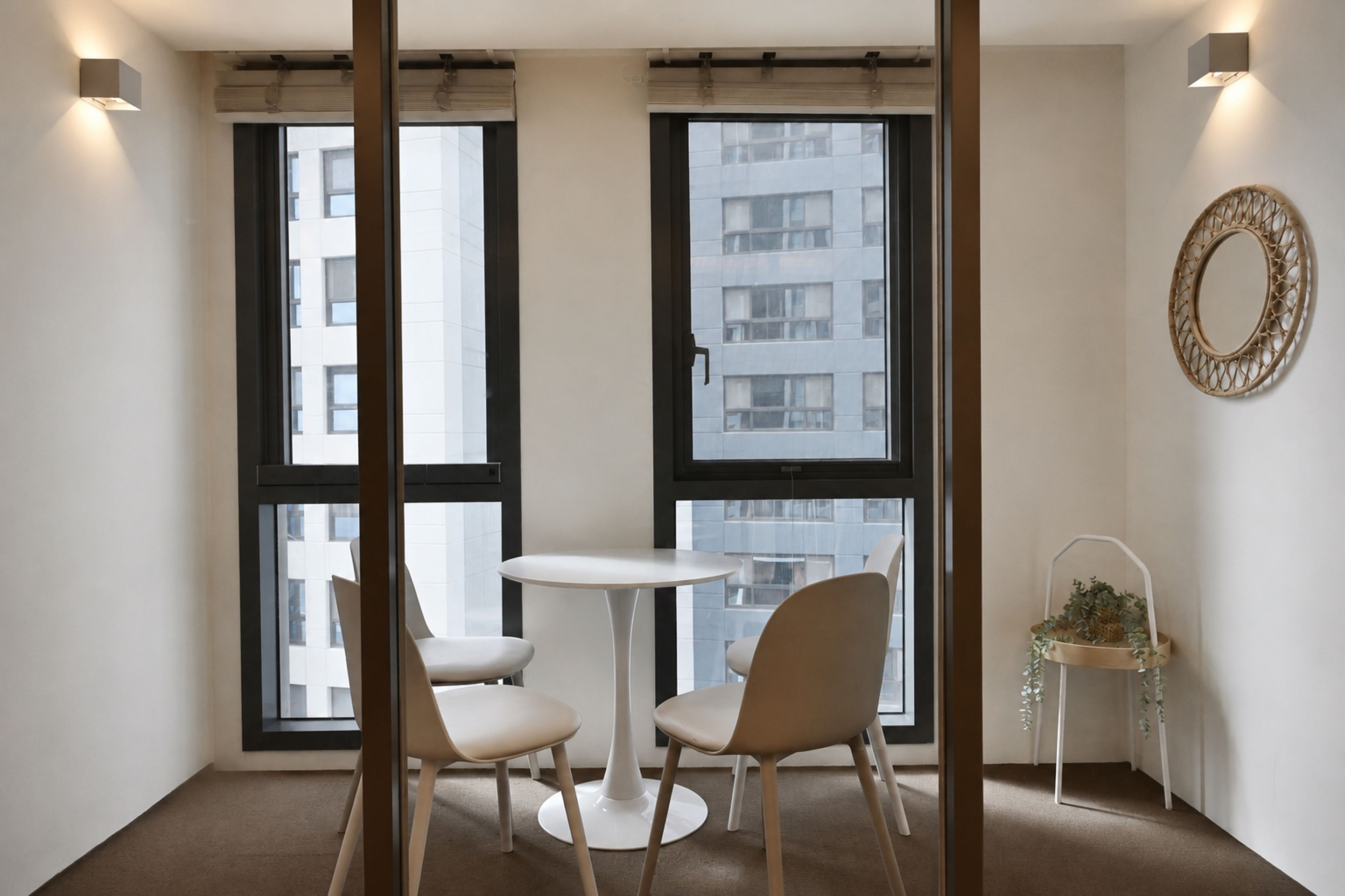 Una habitación moderna con dos sillas alrededor de una mesita pequeña, ventanas amplias y un espejo decorativo en la pared.