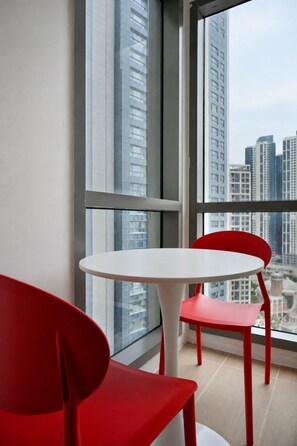 A modern room with a white round table and two red chairs, large windows offering a view of high-rise buildings.