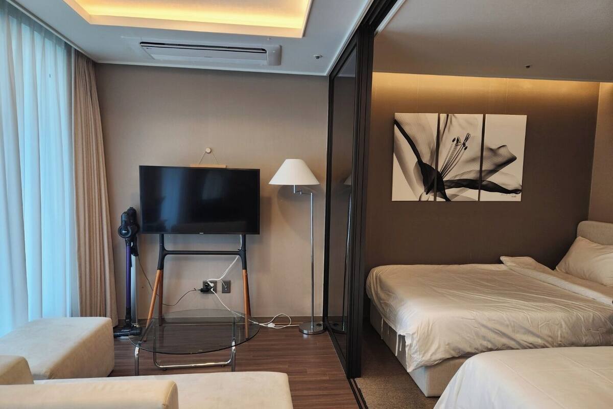 Una habitación de hotel moderna con un televisor de pantalla plana sobre un soporte, una mesa de centro de vidrio, un sofá, una cama y un cuadro en la pared.