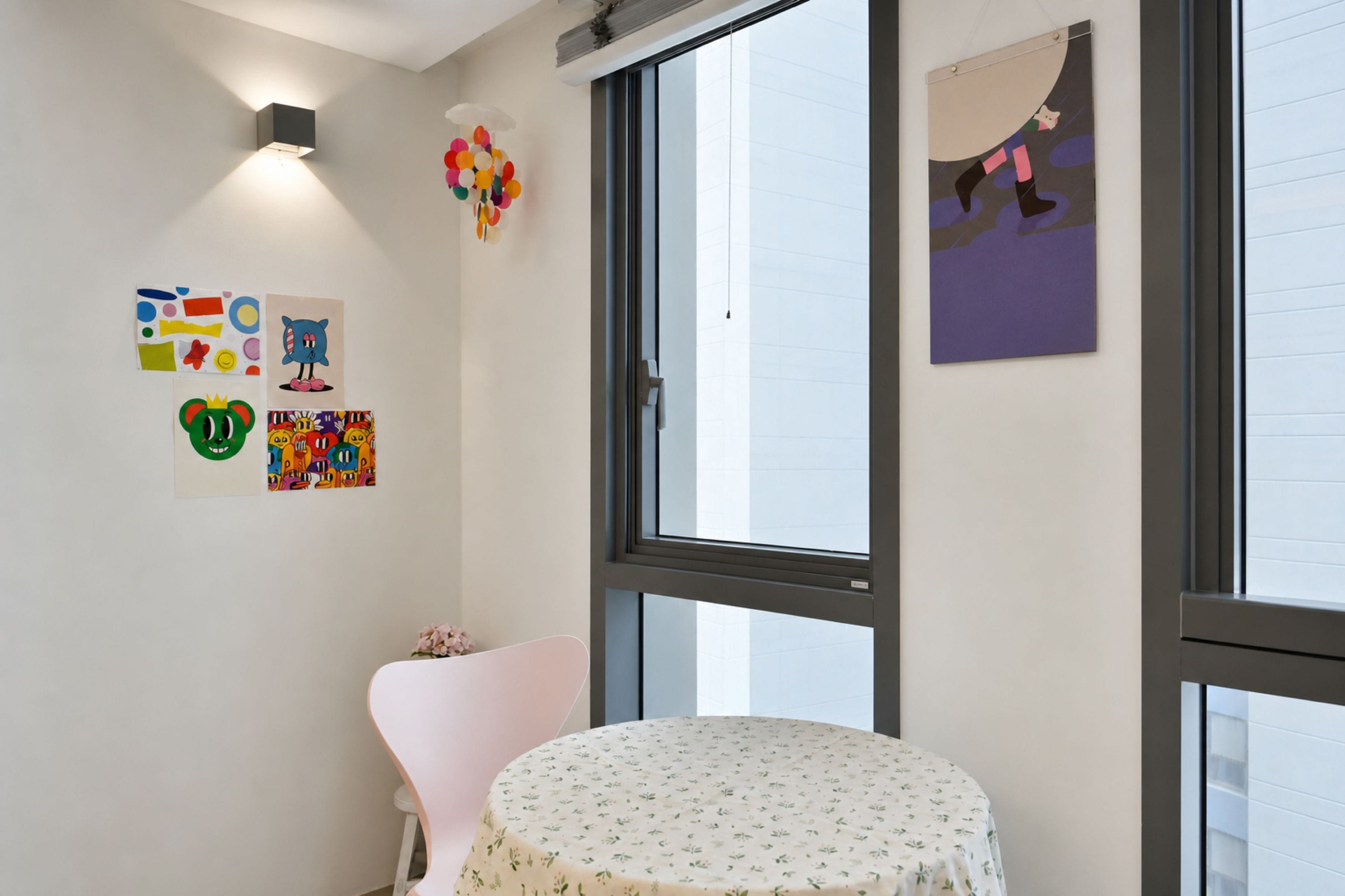A room with a large window, a round table with a floral tablecloth, and various children's artwork on the walls.
