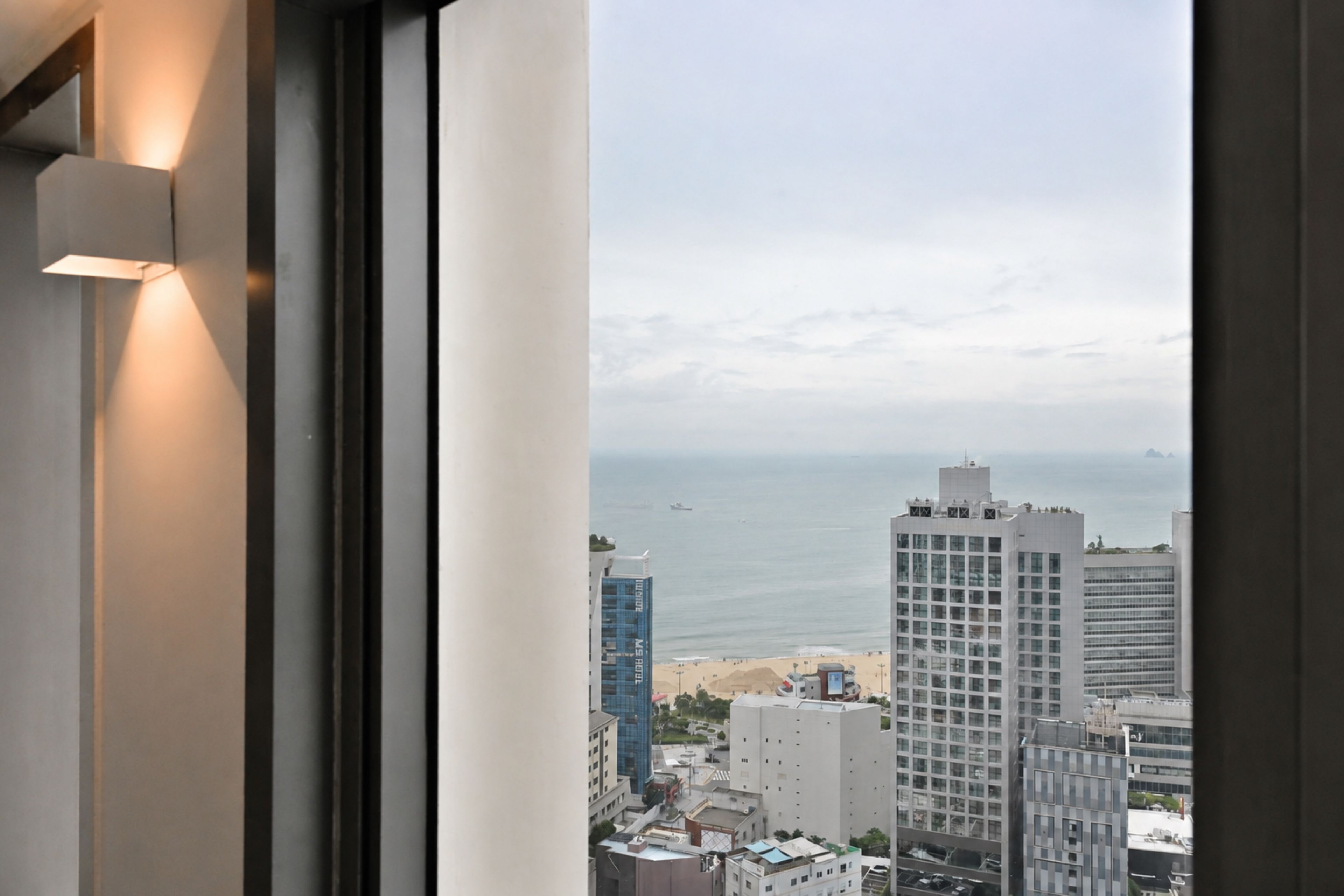 A view of a coastal cityscape through a window, featuring high-rise buildings, a beach, and the ocean.