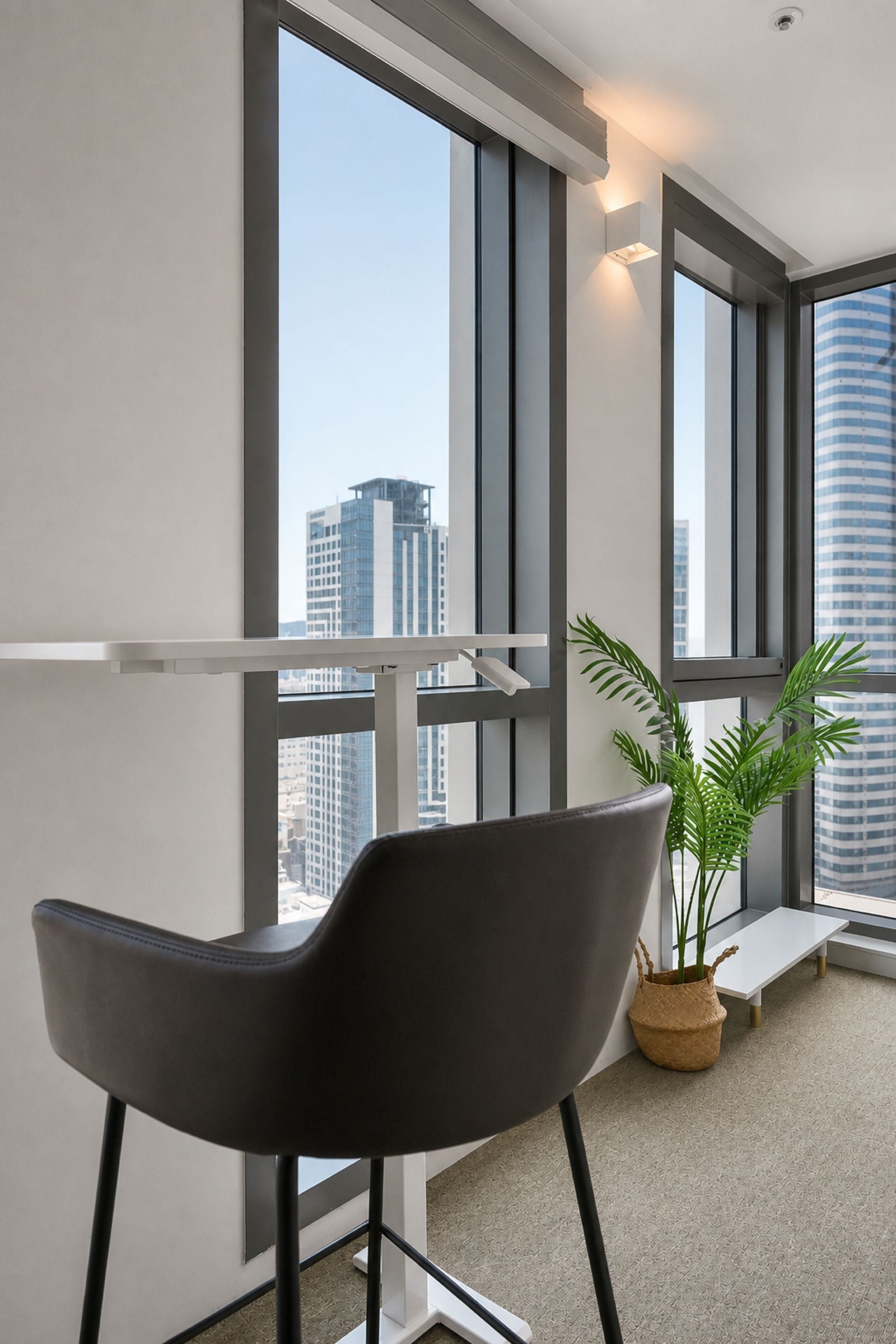 Un espacio de oficina moderno con una silla de escritorio negra, una planta en maceta y vistas de un paisaje urbano a través de grandes ventanales.