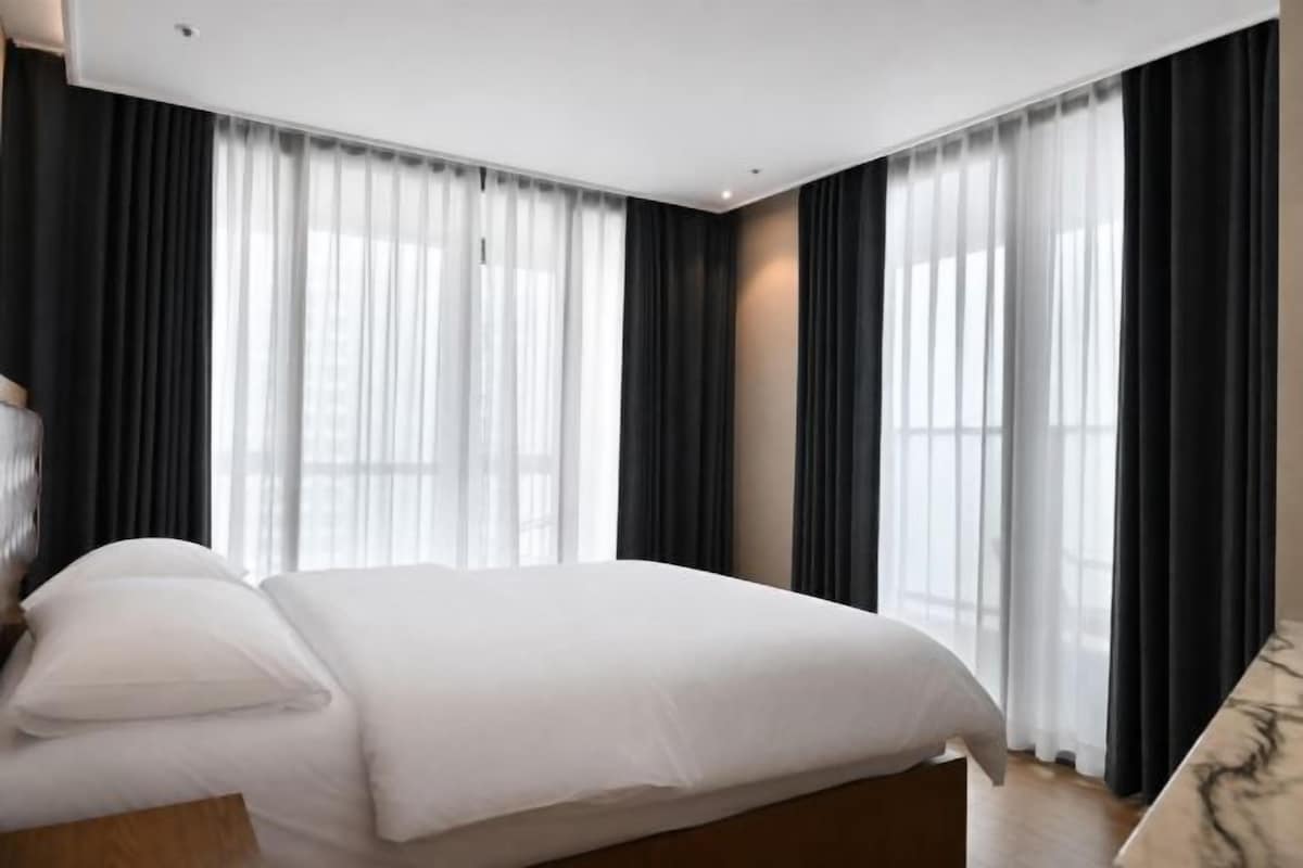 Habitación de hotel con una cama, cabecera de madera y un ventanal con cortinas translúcidas.