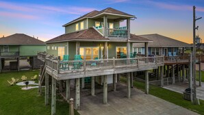 A two-story waterfront house with a deck, outdoor seating, and a view of the water. - Retreat | Hot Tub, Game Room, Firepit & Gulf Views (Bolivar Peninsula)