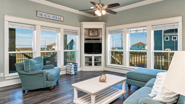 A living room with a view of the beach, a ceiling fan, and a sign that reads "Beach House". - Retreat | Hot Tub, Game Room, Firepit & Gulf Views (Bolivar Peninsula)