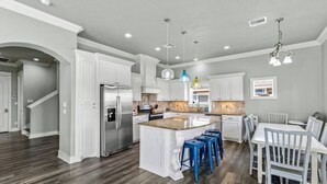 A kitchen with white cabinetry, a central island with blue barstools, and a dining area with white chairs. - Retreat | Hot Tub, Game Room, Firepit & Gulf Views (Bolivar Peninsula)