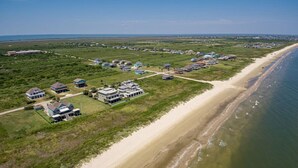 An aerial view of a coastal residential area with houses along the beach, a large building with a prominent roof, and a clear view of the ocean. - Retreat | Hot Tub, Game Room, Firepit & Gulf Views (Bolivar Peninsula)