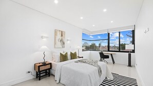 Un dormitorio moderno con una cama grande, un escritorio y vistas de la ciudad a través de un ventanal amplio.