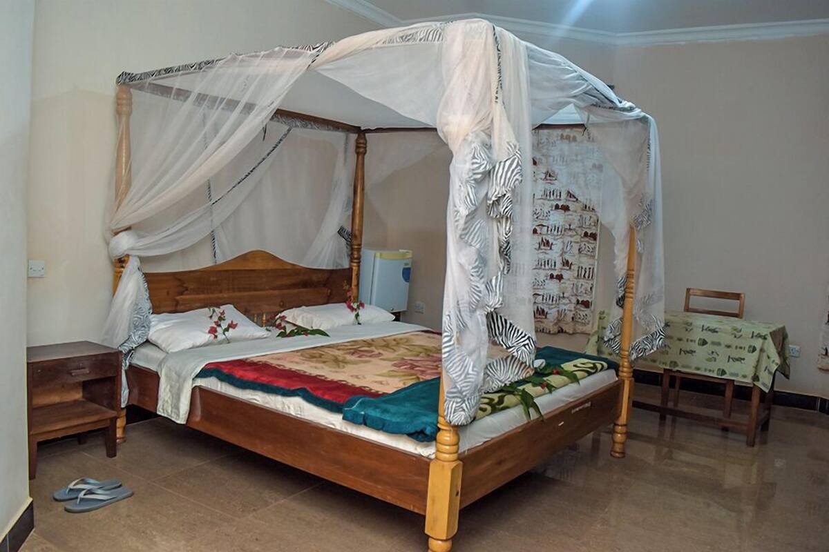 A four-poster bed with a canopy, a wooden headboard, and a floral bedspread. There is a small wooden nightstand to the left of the bed and a chair with a table to the right.