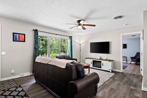 A modern living room with a dark leather sofa, a ceiling fan, a flat-screen TV, and a large window with curtains. - Belle Maison Intermediate Lake Cottage (Bellaire)