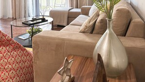 A living room with a beige sofa, a glass coffee table, a wooden side table, a vase with a plant, and a hanging light fixture.