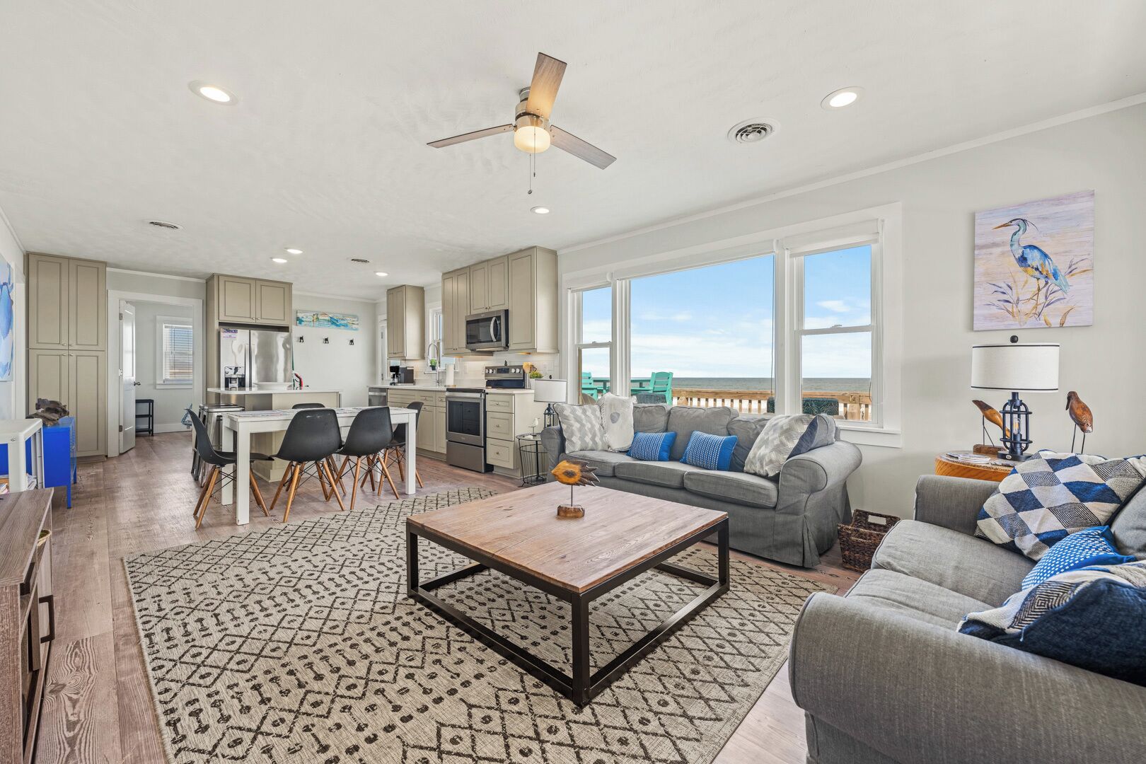 A spacious living area with a large window overlooking the ocean, a dining area with a wooden table, and a kitchen with modern appliances.