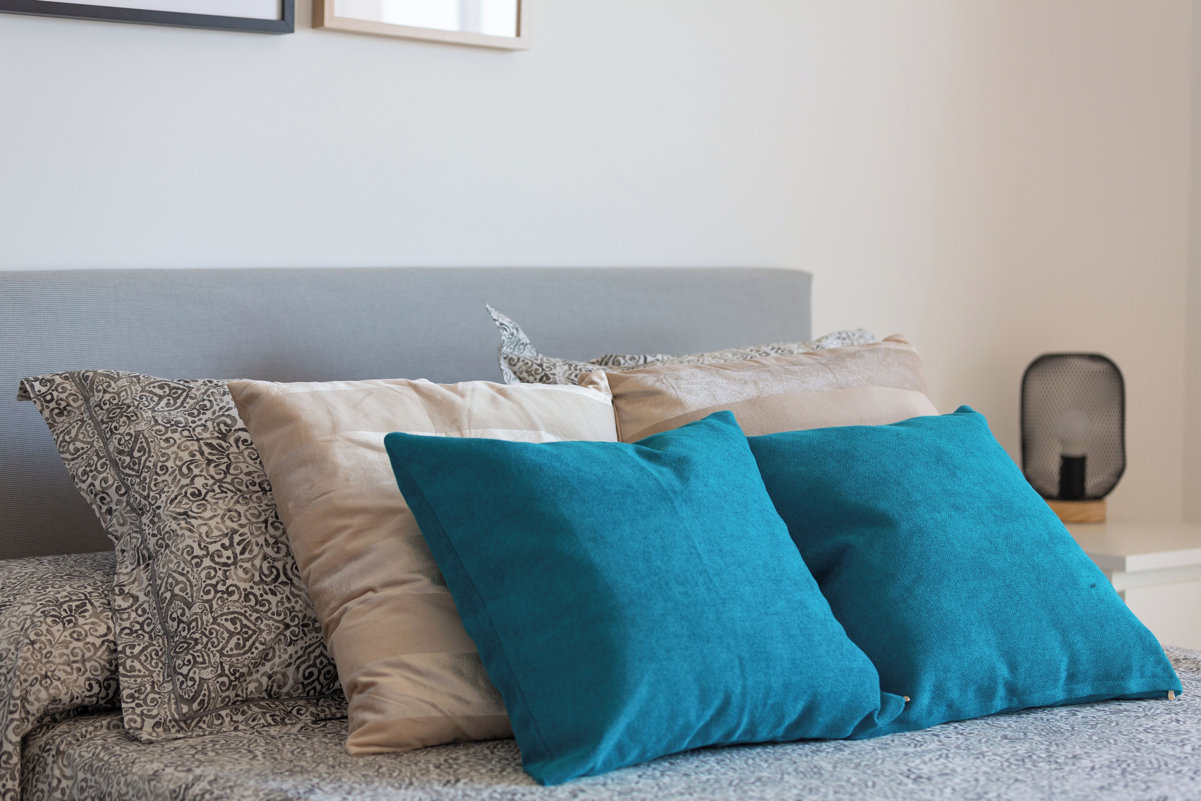 A bed with patterned and solid-colored pillows, a grey headboard, and a small table with a mesh screen.