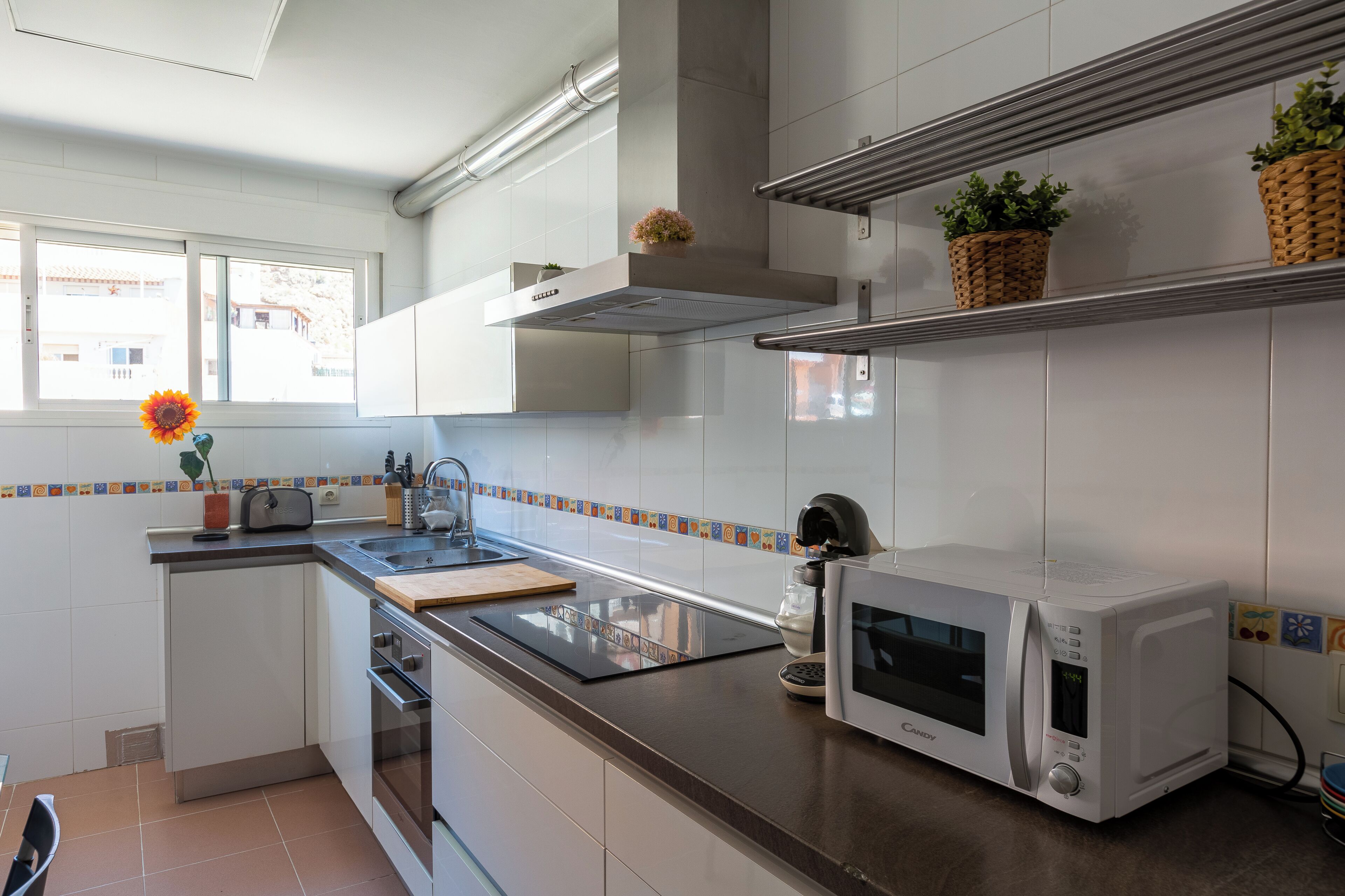 A modern kitchen with white cabinets, a stainless steel range hood, a microwave, and a window with a view of buildings outside.