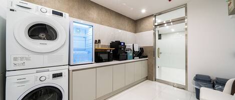 A modern laundry room with stacked washers and dryers, a coffee machine, and a glass door leading to a shower area.